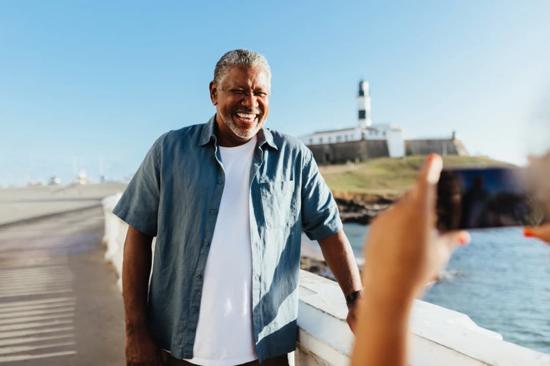 Homem aposentado pelo INSS sorrindo na praia, usando camisa azul por cima de uma camiseta branca.