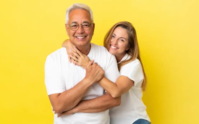 Casal sorrindo após conquistar seu auxílio no INSS. Ambos usam camisetas brancas em um fundo amarelo.